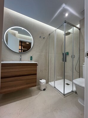 Een moderne badkamer met een houten wastafelmeubel, een ronde spiegel, een douchecabine met glazen wanden en een toilet.