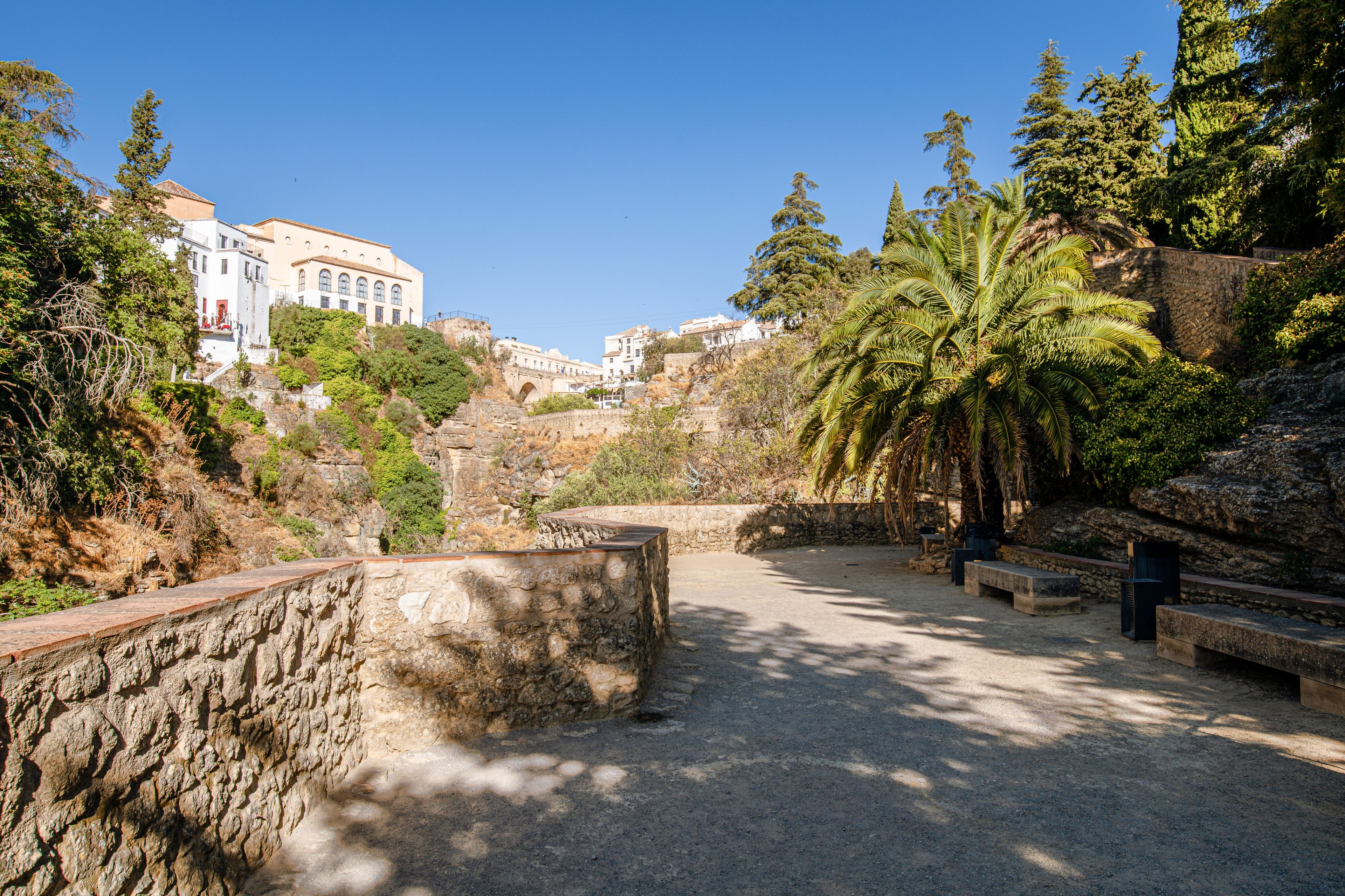 A paved area with stone walls and benches, surrounded by greenery and buildings in the background.