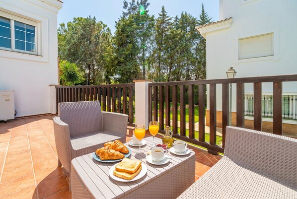 Eine Terrasse mit Korbmöbeln, Frühstücksgebäck und Blick auf Bäume und ein Gebäude.