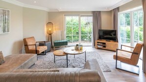 Een moderne woonkamer met een bank, salontafel en televisie, grote ramen en uitzicht op groen.