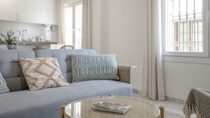 A modern living room with a grey sofa, patterned cushions, a glass coffee table, and a view of a balcony with white railings.