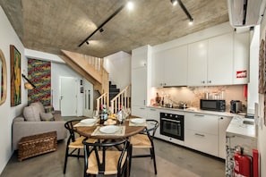 A modern kitchen with white cabinets, a dining area with a wooden table and chairs, and a staircase leading to another level. - Cozy Mozambican Retreat (Cabanas de tavira)