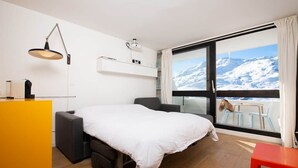 Ein Hotelzimmer mit Bett, Schreibtisch, Stuhl, kleinem Tisch, Fenster mit Blick auf schneebedeckte Berge und einem Balkon mit einem Hund.