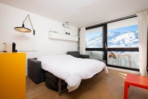 A hotel room with a bed, a desk, a chair, a small table, a window with a view of snow-covered mountains, and a balcony with a dog.