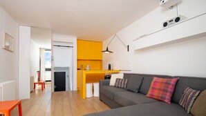 Ein modernes Wohnzimmer mit einem grauen Sofa, einem kleinen orangefarbenen Tisch und einer Küchenzeile mit gelben Schränken.