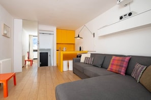 A modern living room with a grey sofa, a small orange table, and a kitchen area with yellow cabinets.