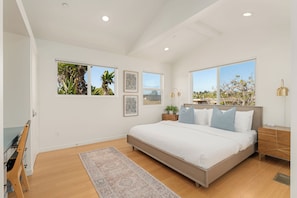 A bedroom with a large bed, a nightstand, a bedside table, a lamp, and a window with a view of trees. - Family Beach House w/ Pool & AC + Walking Distance to Shops + Food (Encinitas)