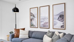 Een moderne woonkamer met een grijze bank, drie ingelijste foto’s van kustlandschappen, een zwarte hanglamp en een houten bijzettafeltje met een plant.