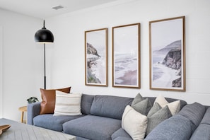 Ein modernes Wohnzimmer mit einem grauen Sofa, drei gerahmten Bildern von Küstenlandschaften, einer schwarzen Pendelleuchte und einem Holztisch mit einer Pflanze.