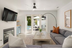 A modern living room with a flat-screen TV, a grey sofa, a coffee table, and a ceiling fan. - Family Beach House w/ Pool & AC + Walking Distance to Shops + Food (Encinitas)