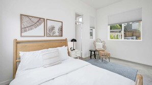 Een slaapkamer met een houten bed, witte beddengoed, een klein nachtkastje met een lamp, een rieten stoel en een raam met uitzicht op groen.