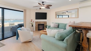Una sala de estar moderna con chimenea, ventilador de techo, televisor grande y vista al mar.