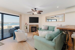 A modern living room with a fireplace, a ceiling fan, a large TV, and a view of the sea. - Ground Floor Waterfront | AC | Private Patio (San Diego)