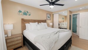 Un dormitorio con cama, mesitas de noche, ventilador de techo y baño visible a través de una puerta abierta.