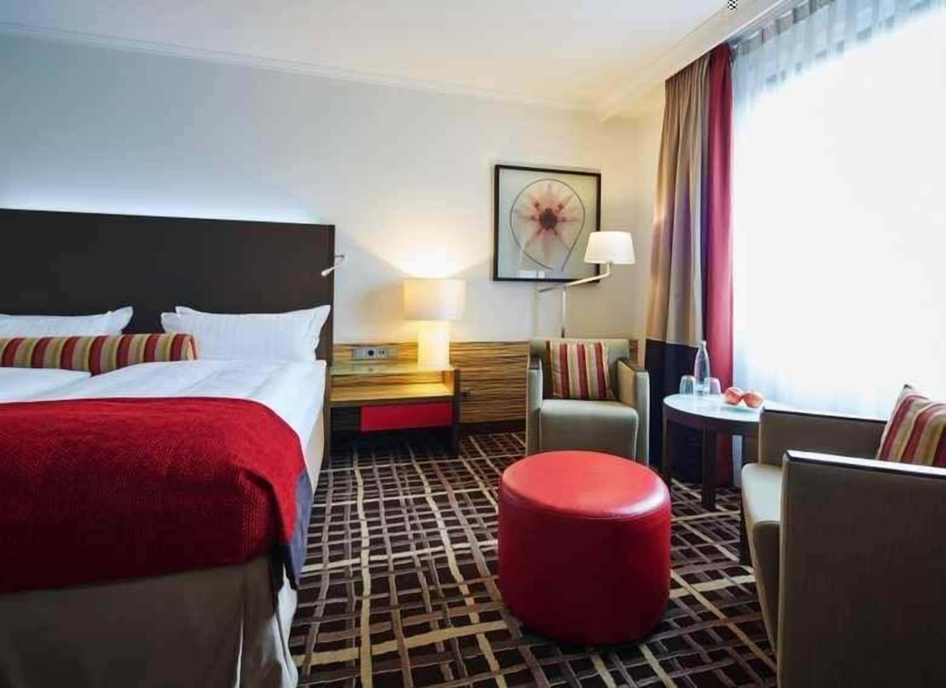 Een hotelkamer met twee bedden, een rode poef, een klein tafeltje met een lamp en een groot raam met gordijnen.