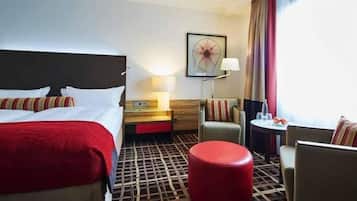 Een hotelkamer met twee bedden, een rode poef, een klein tafeltje met een lamp en een groot raam met gordijnen.