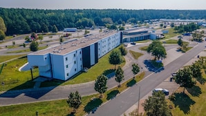Um edifício com vários andares e telhado plano, rodeado por um estacionamento e áreas verdes.