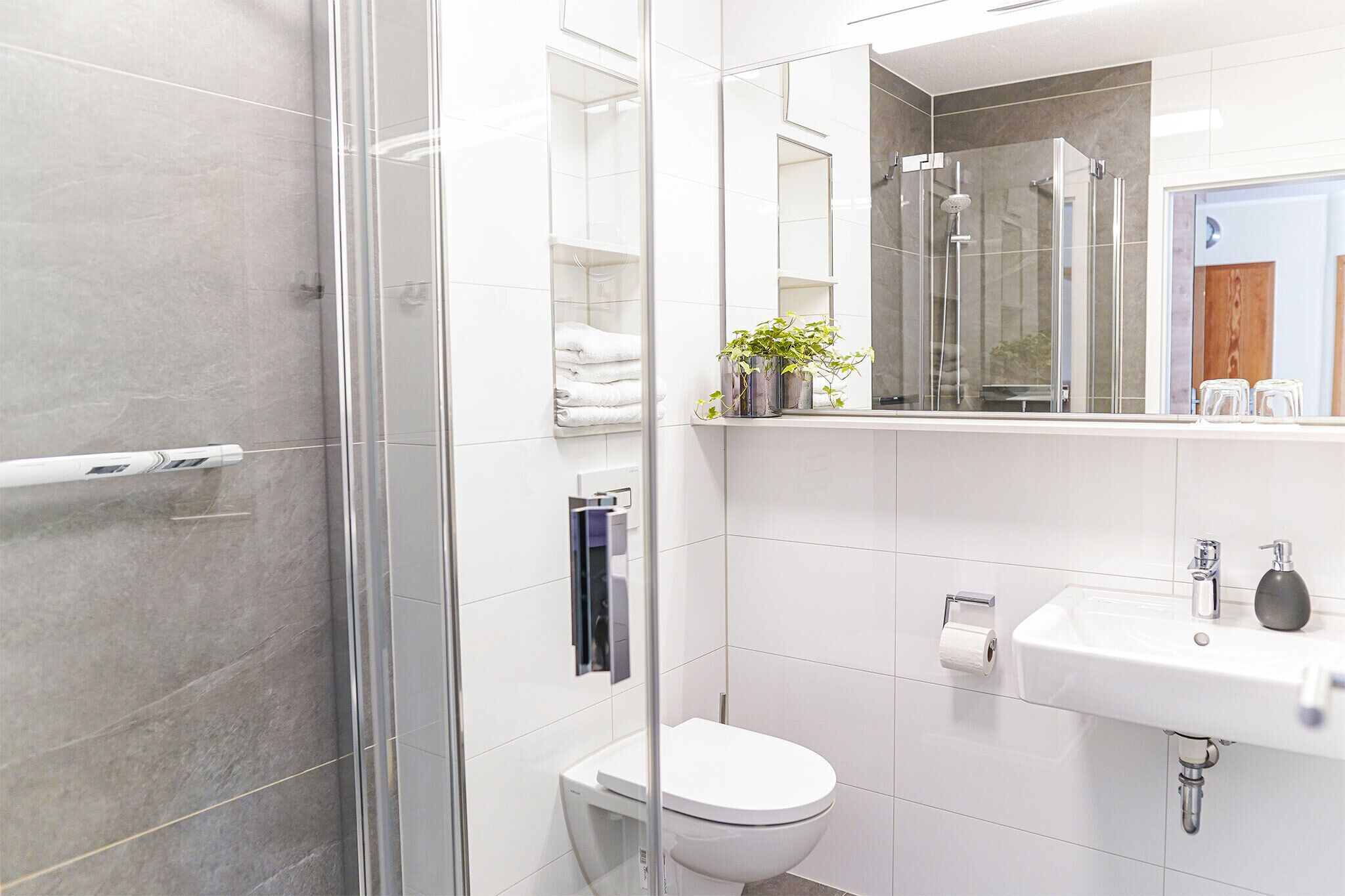 Ein modernes Badezimmer mit Duschkabine aus Glas, einer weißen Toilette, einem Waschbecken mit Spiegel und einem Regal mit Handtüchern und einer Pflanze.