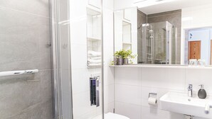 Moderni kylpyhuone, jossa on lasinen suihkuseinä, valkoinen wc-istuin, peilillä varustettu pesuallas sekä hylly, jossa on pyyhkeitä ja kasvi.