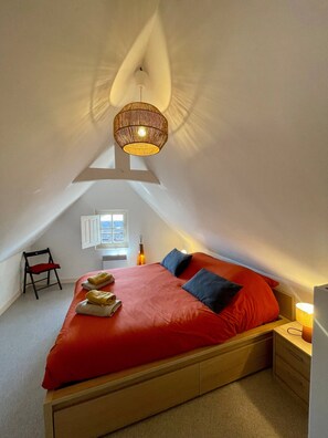 Een slaapkamer met een schuine kap, een bed met rode beddengoed, een kleine stoel, een nachtkastje met een lamp en een raam met jaloezieën.