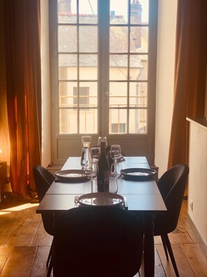 Una mesa de comedor preparada para cuatro personas, sillas, una ventana con vista a edificios y cortinas a los lados.