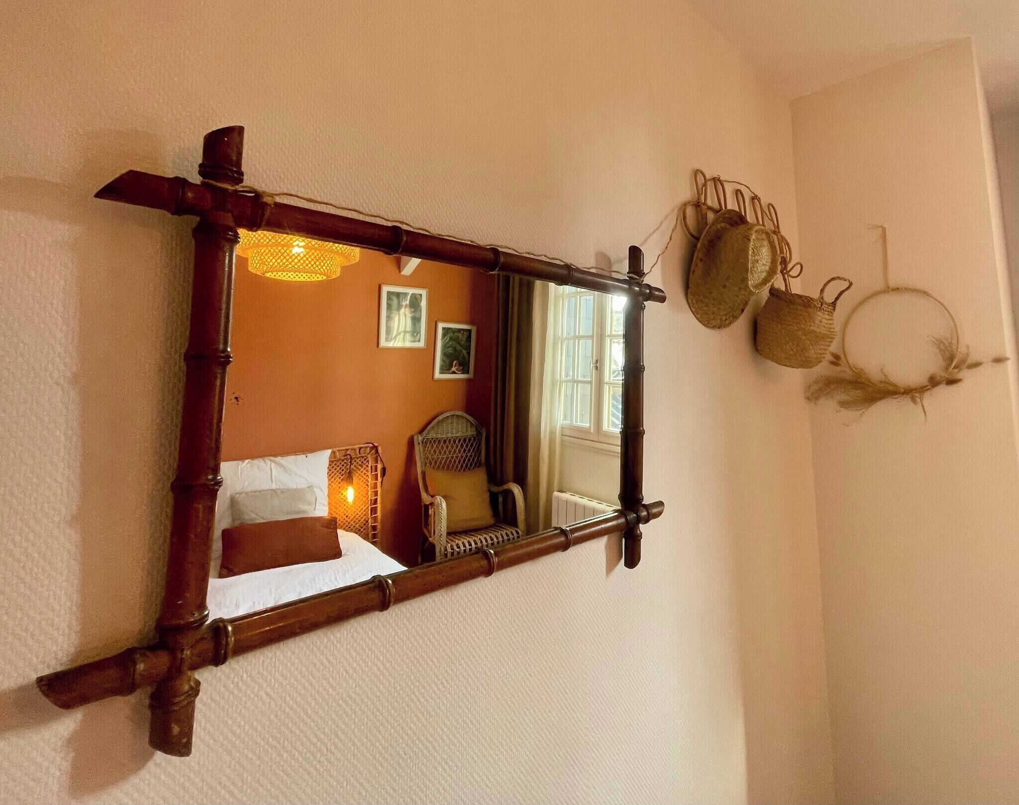 Um espelho com moldura de madeira, uma cama com lençóis brancos, uma cadeira de vime e uma janela com cortinas.