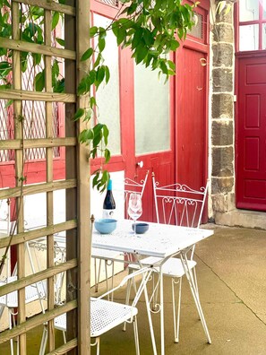 Un juego de mesa y sillas blancas al aire libre, frente a un edificio con puertas rojas y un muro de piedra.