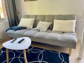 Ein graues Sofa mit vielen Kissen, ein kleiner weißer Tisch mit einer Fernbedienung und ein dunkelblauer Teppich mit weißen Mustern.