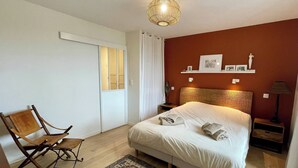 A bedroom with a bed, a chair, a bedside table, a lamp, a rug, and a wall-mounted shelf with pictures.