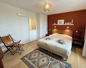 Ein Schlafzimmer mit Bett, Stuhl, Nachttisch, Lampe, Teppich und einem an der Wand befestigten Regal mit Bildern.
