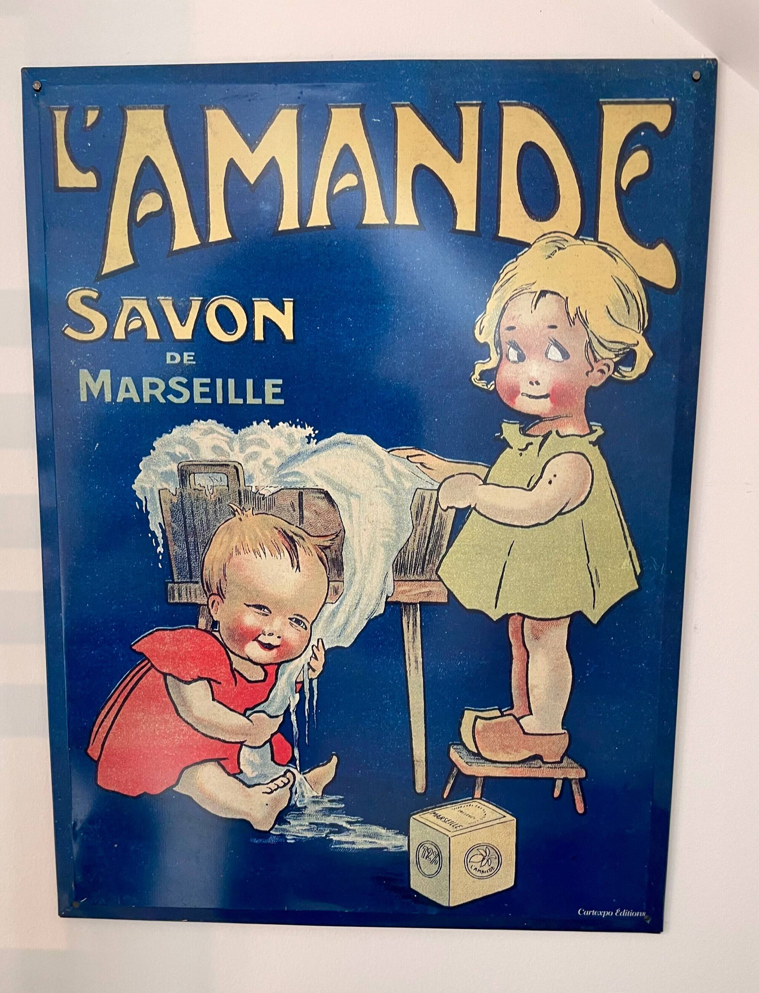 Une affiche ancienne intitulée « L’AMANDE, SAVON DE MARSEILLE », illustrée de deux enfants en train de laver du linge.