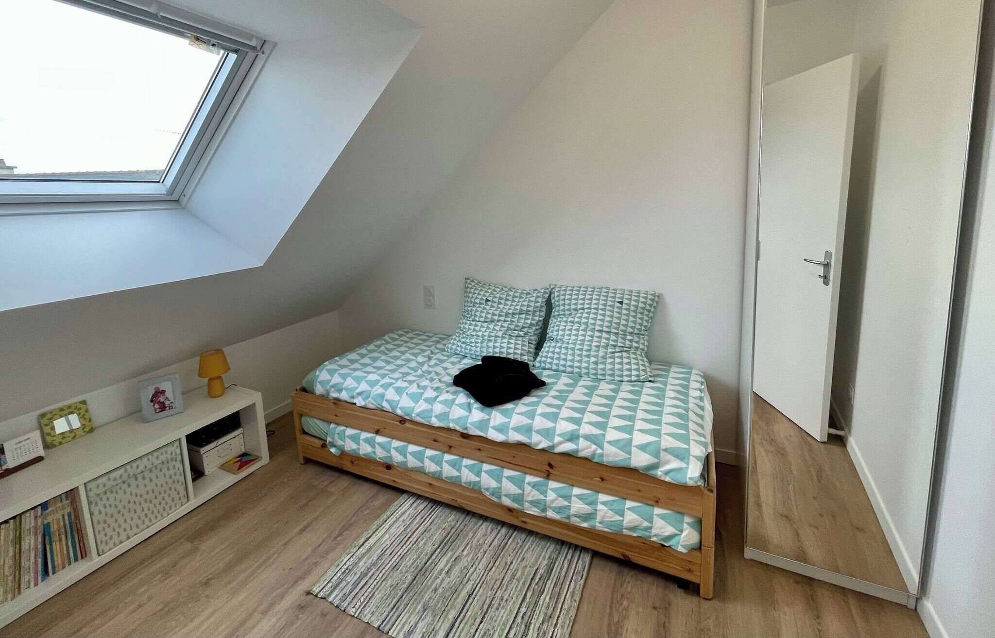Une chambre avec un plafond en pente, un puits de lumière, un lit avec une literie à motifs, une table de chevet avec une lampe et une bibliothèque.
