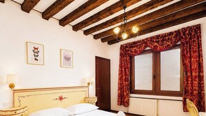 A hotel room with a bed, a nightstand, a lamp, a window with curtains, and a wooden ceiling. - Room-venezia Baroque Charm (Venice)