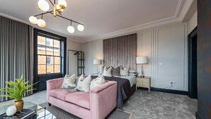 A modern bedroom with a pink sofa, a bed with a headboard, a bedside table, a lamp, and a large window. - One Capel Suites (Dublin)