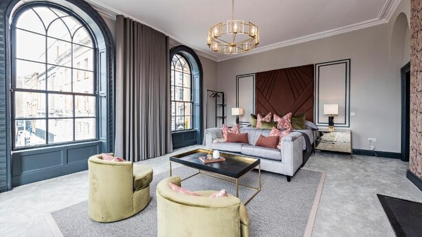 A modern living room with a sofa, ottomans, a coffee table, and a large window with curtains. - One Capel Suites (Dublin)