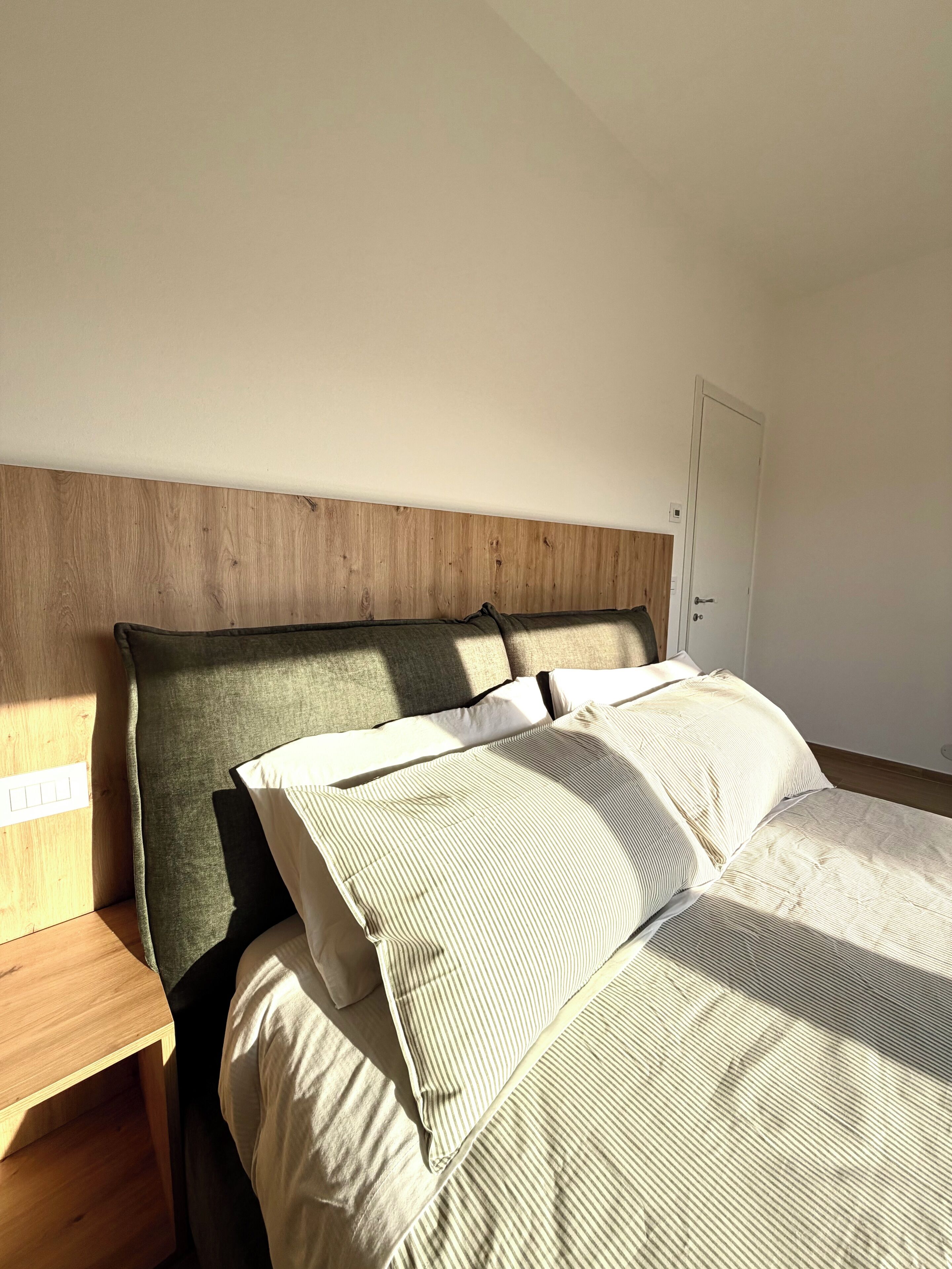Uma cama com cabeceira de madeira, uma mesa de cabeceira e uma porta num quarto com paredes brancas.