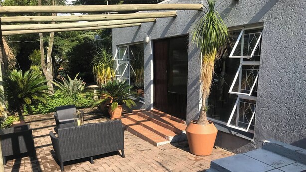 Eine Terrassenfläche mit einer Pergola, Gartenmöbeln, Blumentöpfen und Blick auf ein Gebäude mit großen Fenstern.