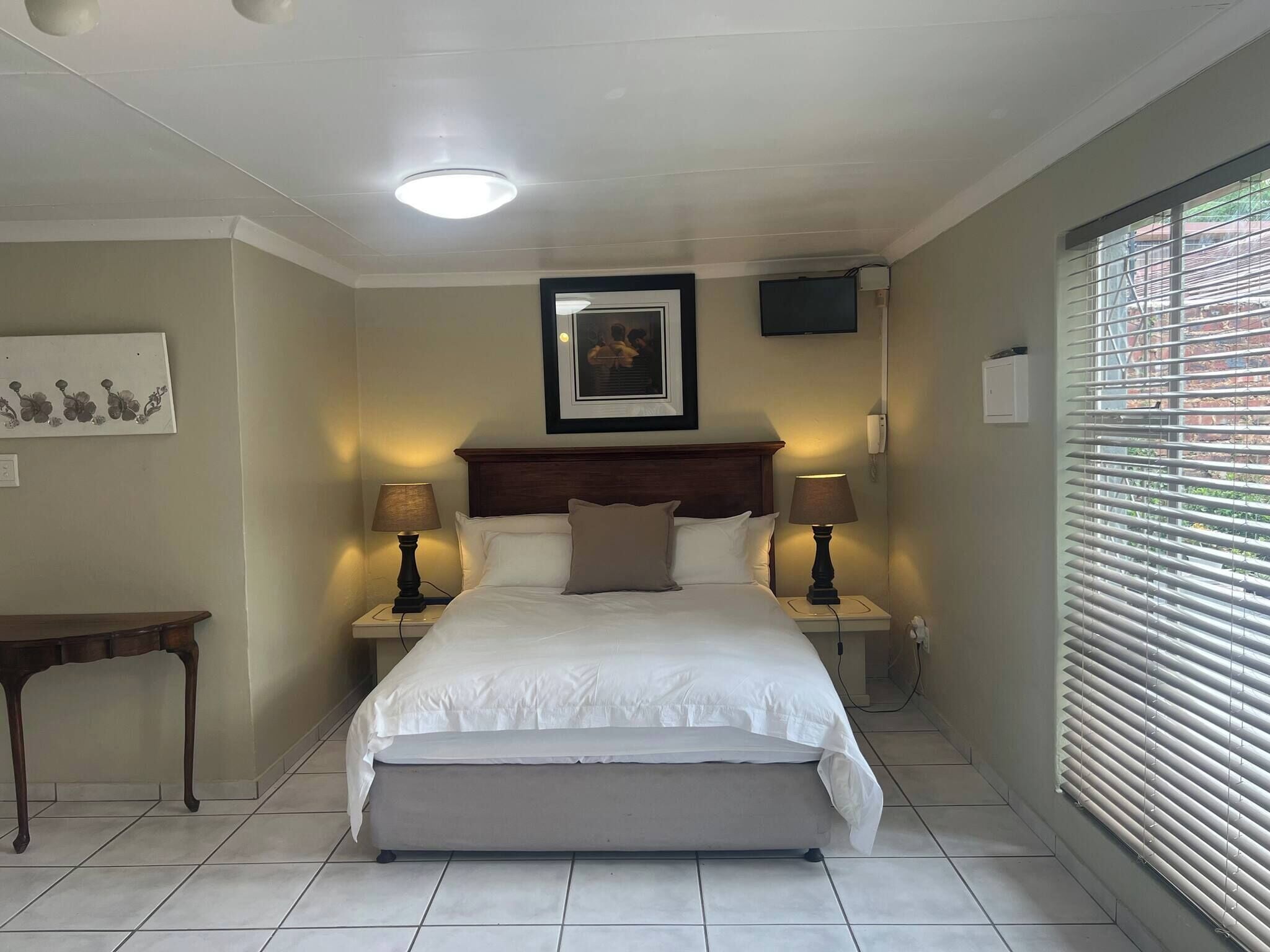 A bedroom with a bed, bedside lamps, a nightstand, a bench, a framed picture, a ceiling light, and a window with blinds.