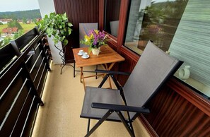 En balkong med ett träbord, två stolar, en krukväxt och en vas med blommor.