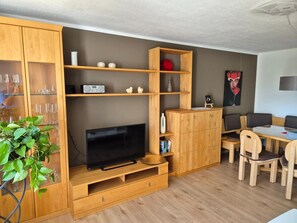 Ein modernes Wohnzimmer mit Flachbildfernseher, Holzschränken, einer Essecke mit Tisch und Stühlen sowie einer Topfpflanze.