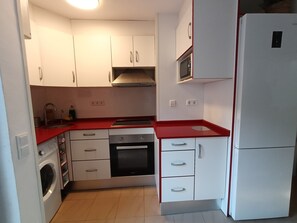 Une cuisine compacte avec des meubles blancs, un plan de travail rouge, un four encastré, un four à micro-ondes et un lave-linge.