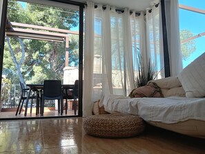 Ein Zimmer mit einer Couch, einem gewebten Hocker, einem Esstisch mit Stühlen und Blick auf Bäume durch große Fenster.