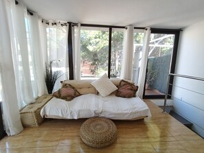 Ein Schlafzimmer mit einem Bett, einem gewebten Hocker und Blick ins Grüne durch große Fenster.