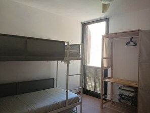 Un lit superposé avec un lit inférieur, une étagère en bois avec une tringle pour suspendre des vêtements, et une fenêtre avec des stores.