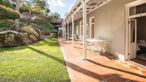 A covered outdoor patio with tables and chairs, a grassy area with rocks, and a building with a tiled roof. - Nantes-Vue (Paarl)