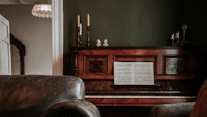 A piano with sheet music, a dark wooden cabinet, and two leather chairs.