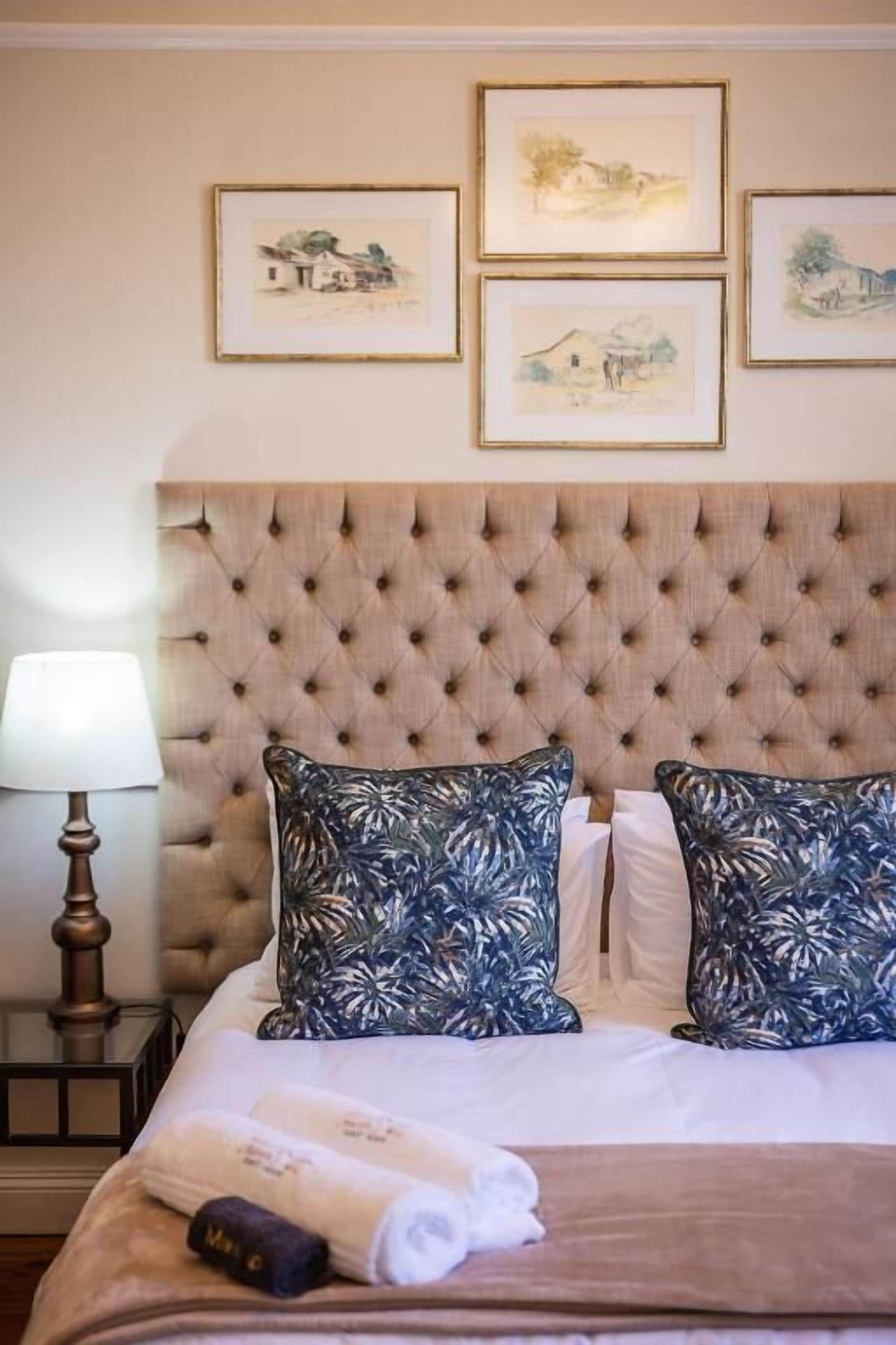 A bed with a tufted headboard, two patterned pillows, a rolled towel, and a bedside lamp.
