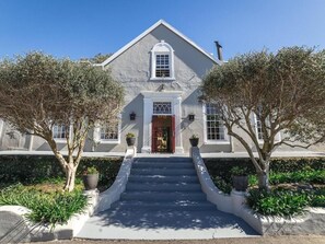 A two-story building with a symmetrical facade, a central entrance with a red door, and a staircase leading to the entrance. - Nantes-Vue (Paarl)