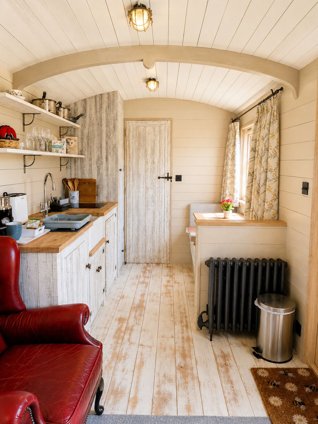 A compact kitchen with wooden cabinets, a red leather chair, a radiator, and a small trash bin.