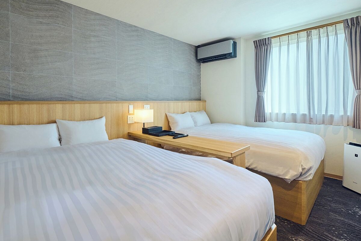 A hotel room with two beds, a bedside table with a lamp, an air conditioning unit, and a window with curtains.